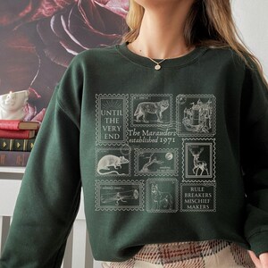 K&ouml;nnte beinhalten: Ein dunkelgr&uuml;ner Pullover mit einem wei&szlig;en Grafikdruck von Briefmarken mit Illustrationen eines Wolfes, einer Ratte, eines Hirsches, eines Baumes, eines Schlosses und eines Hundes. Die Briefmarken tragen den Text "The Marauders established 1971", "Until the very end", "I solemnly swear", "Mischief managed", "Moony", "Wormtail", "Padfoot", "Prongs" und "Rule breakers mischief makers".