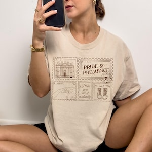 Op de afbeelding: Beige t-shirt met een bruin stempelontwerp. Het ontwerp bevat de woorden "Pride & Prejudice" en illustraties van een gebouw, handen en laarzen. De tekst "I love you most ardently" is ook opgenomen.