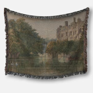 May include: A woven throw blanket depicting a landscape painting of a castle on a lake with trees and swans. The artwork is in muted tones of green, brown, and beige. The blanket has a fringed edge.
