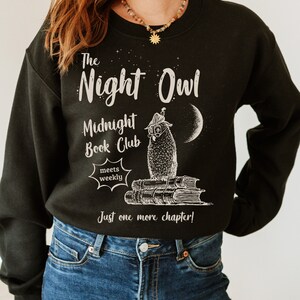 May include: Black sweatshirt with a white graphic of an owl wearing glasses and a hat, sitting on a stack of books. The text reads "The Night Owl Midnight Book Club meets weekly Just one more chapter!"