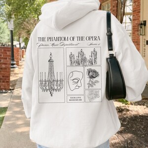 May include: White hoodie featuring a graphic design from "The Phantom of the Opera." The design includes a chandelier, candles, a mask, a rose, and the text "YOUR LOVE REDEEMS ME."