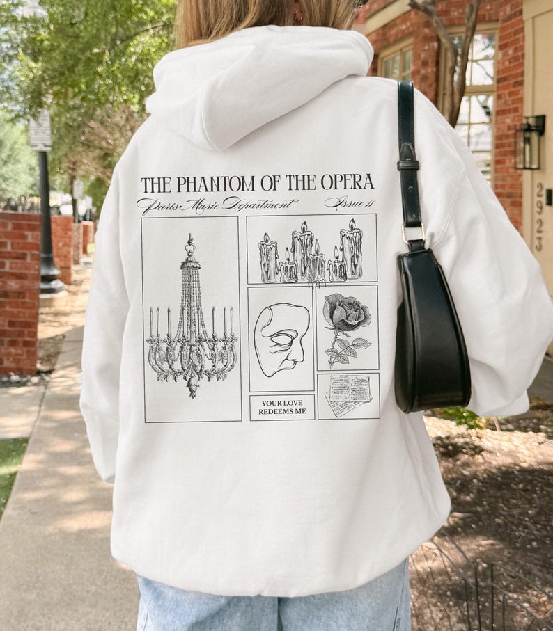 May include: White hoodie featuring a graphic design from "The Phantom of the Opera." The design includes a chandelier, candles, a mask, a rose, and the text "YOUR LOVE REDEEMS ME."