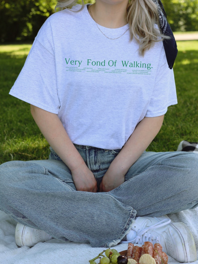 May include: A person wearing a grey t-shirt with the text "Very Fond Of Walking." in green lettering. The person is sitting on a white blanket with their legs crossed. They are wearing blue jeans and white sneakers.
