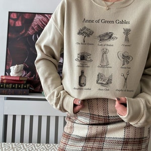 Op de afbeelding: Een beige sweatshirt met een grafische print van Anne of Green Gables en verschillende items uit het boek. De tekst op het sweatshirt luidt "Anne of Green Gables" en bevat de namen van items uit het boek, zoals "De Sneeuwkoningin", "Lady of Shalott", "Wortelen", "Vanilletaart", "Puffmouwen", "Strohoed", "Frambozenlikeur", "Verhalenclub" en "Diepten van wanhoop".