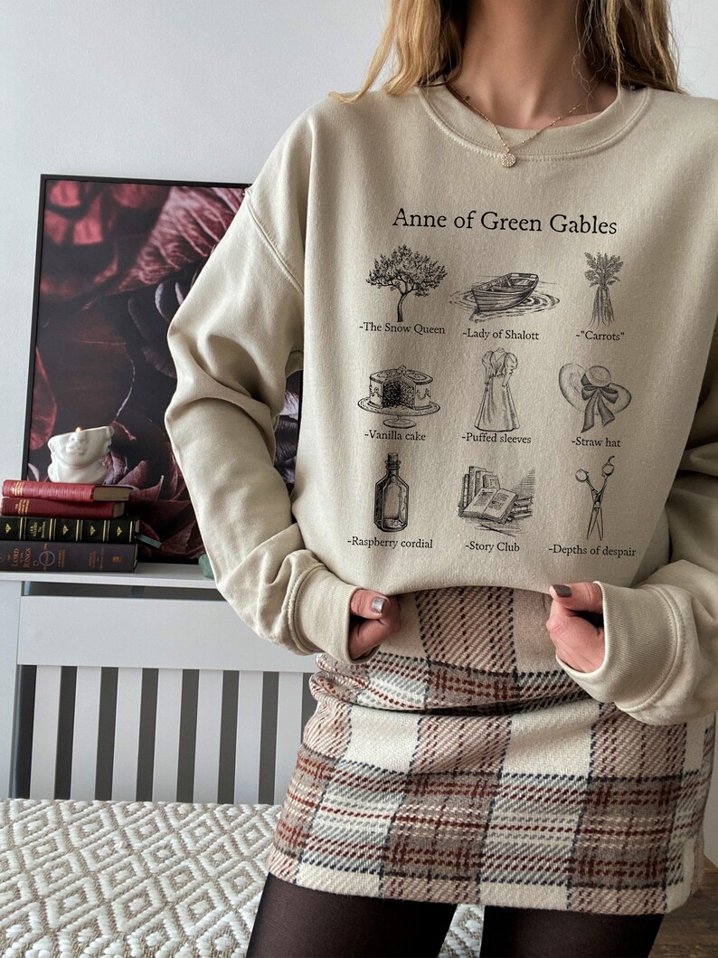 Op de afbeelding: Een beige sweatshirt met een grafische print van Anne of Green Gables en verschillende items uit het boek. De tekst op het sweatshirt luidt "Anne of Green Gables" en bevat de namen van items uit het boek, zoals "De Sneeuwkoningin", "Lady of Shalott", "Wortelen", "Vanilletaart", "Puffmouwen", "Strohoed", "Frambozenlikeur", "Verhalenclub" en "Diepten van wanhoop".