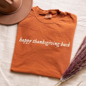 Puede incluir: Una camiseta naranja con la frase "happy thanksgiving back" en escritura blanca. Un sombrero marrón y un ramo de flores secas también están en la imagen. La camiseta está doblada y tiene una etiqueta en el cuello.