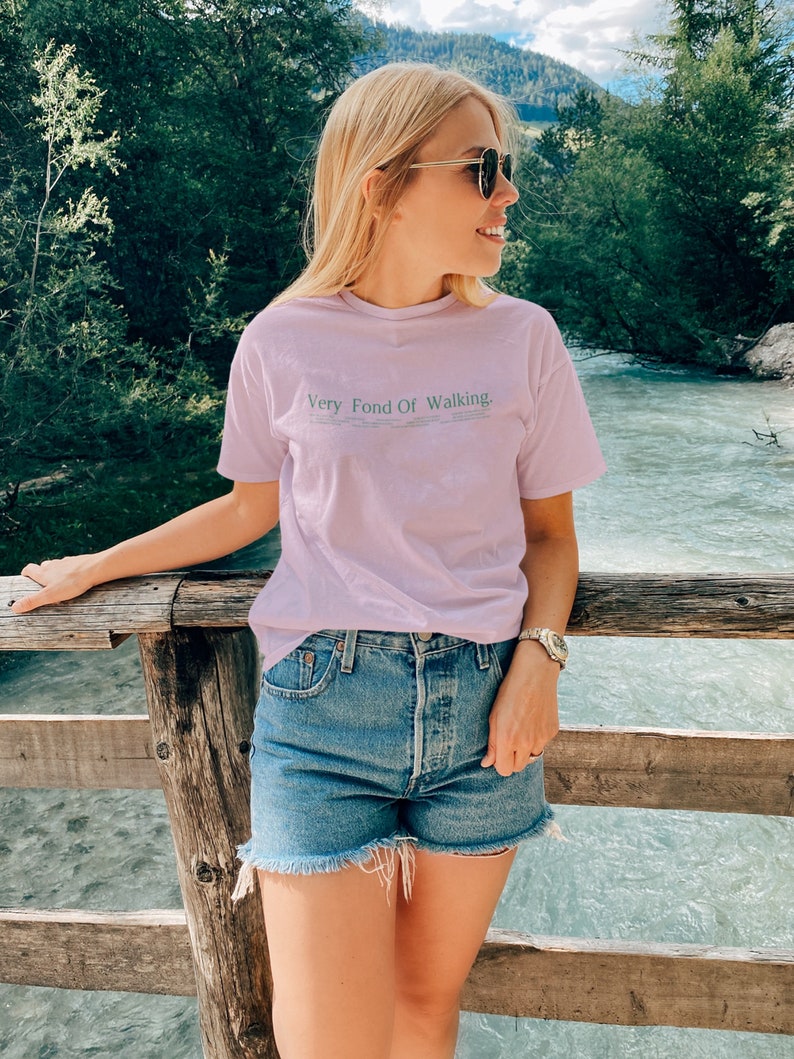 May include: A woman wearing a light purple t-shirt with the text "Very Fond Of Walking" in green lettering. She is wearing denim shorts and sunglasses and is standing on a wooden bridge.