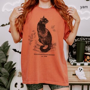 Op de afbeelding: Verbrand oranje t-shirt met een illustratie van een zwarte kat die op een stapel boeken en vallende bladeren zit. De tekst luidt "Halloweentown Books est. 1998". Het shirt heeft een ronde hals en korte mouwen.