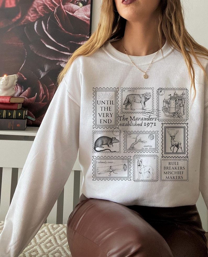 K&ouml;nnte beinhalten: Wei&szlig;er Pullover mit einem grafischen Druck von Briefmarken mit Illustrationen eines Wolfes, einer Burg, einer Ratte, eines Baumes, eines Hirsches, eines aufgerollten Pergaments und eines Hundes. Die Briefmarken tragen den Text "The Marauders established 1971" und "Rule Breakers Mischief Makers".