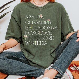 May include: A green t-shirt with a white graphic print of the words "Azalea", "Oleander", "Belladonna", "Foxglove", "Hellebore", and "Wisteria".