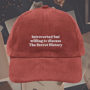 May include: Red corduroy baseball cap with white embroidered text that reads "introverted but willing to discuss The Secret History".