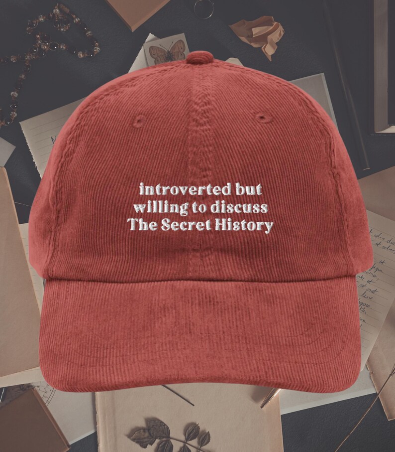 May include: Red corduroy baseball cap with white embroidered text that reads "introverted but willing to discuss The Secret History".