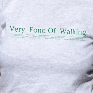 May include: Gray t-shirt with the text "Very Fond Of Walking." in green. The text is surrounded by a list of phrases in green, including "Cross field after field", "Jump over styles", "Spring over puddles", "Observe the glories of nature", "Bewitch men body & soul", "Six inches deep in mud", "Admire the natural beauty", "Take a turn about the room", "Discuss secret affairs", "Delight in anything ridiculous", "Glow with the warmth of exercise", "Bio adieu to disappointment", and "Distance is nothing when one has a motive."