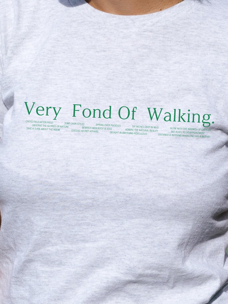 May include: Gray t-shirt with the text "Very Fond Of Walking." in green. The text is surrounded by a list of phrases in green, including "Cross field after field", "Jump over styles", "Spring over puddles", "Observe the glories of nature", "Bewitch men body & soul", "Six inches deep in mud", "Admire the natural beauty", "Take a turn about the room", "Discuss secret affairs", "Delight in anything ridiculous", "Glow with the warmth of exercise", "Bio adieu to disappointment", and "Distance is nothing when one has a motive."