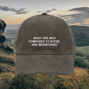 Peut inclure: Une casquette de baseball en velours côtelé vert avec le texte "WHAT ARE MEN COMPARED TO ROCKS AND MOUNTAINS?" brodé en blanc.