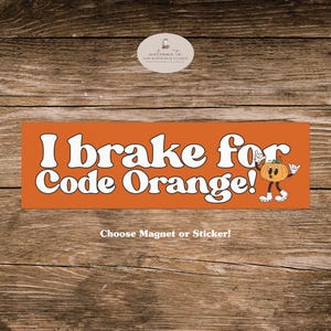 May include: An orange magnet or sticker with the text "I brake for Code Orange!" in white, featuring a cartoon pumpkin. The phrase "Choose Magnet or Sticker!" is below the text. The background is a wooden surface.