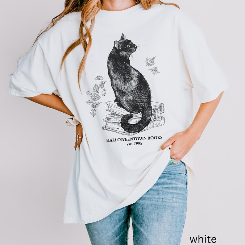 Op de afbeelding: Cr&egrave;mekleurig T-shirt met een illustratie van een zwarte kat op boeken. De afbeelding bevat de tekst "Halloweentown Books est. 1998" en bladeren. Het shirt heeft een losse pasvorm.