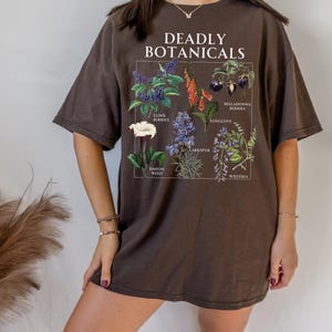 May include: Chocolate-colored t-shirt featuring a botanical illustration with the text "DEADLY BOTANICALS." The graphic includes drawings of various plants such as elderberries, foxglove, and larkspur. The word "chocolate" is printed at the bottom.