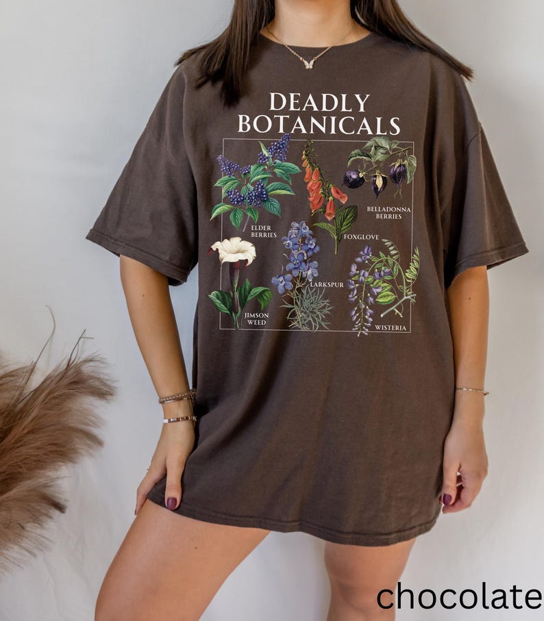 May include: Chocolate-colored t-shirt featuring a botanical illustration with the text "DEADLY BOTANICALS." The graphic includes drawings of various plants such as elderberries, foxglove, and larkspur. The word "chocolate" is printed at the bottom.