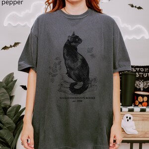 Op de afbeelding: Donkergrijze t-shirt met een illustratie van een zwarte kat die op boeken zit met vallende bladeren. De tekst "Halloweentown Books est. 1998" staat eronder. Het shirt heeft een vintage, licht vervaagde uitstraling.