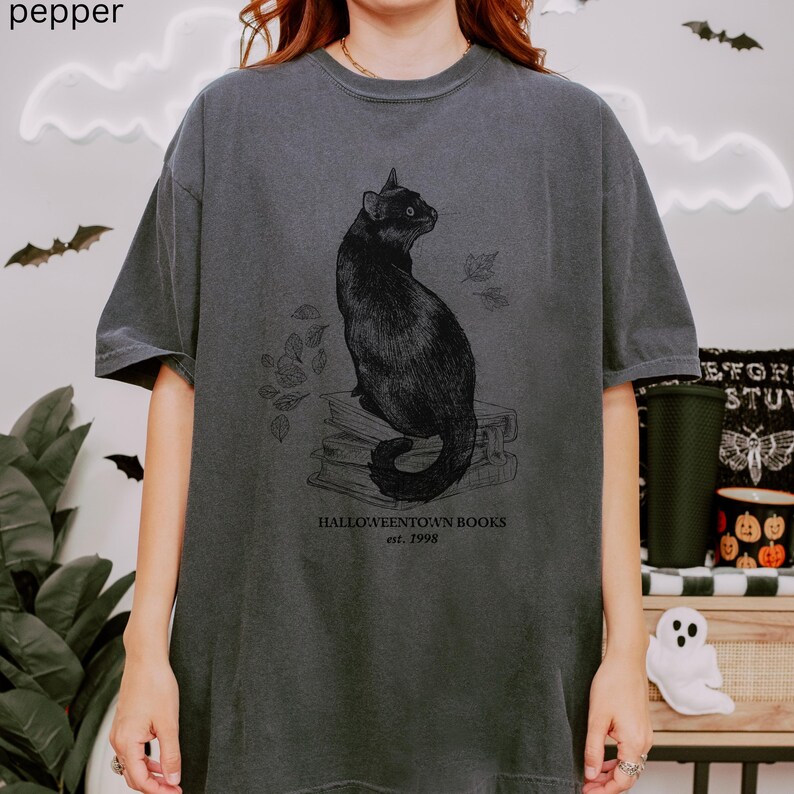Op de afbeelding: Donkergrijze t-shirt met een illustratie van een zwarte kat die op boeken zit met vallende bladeren. De tekst "Halloweentown Books est. 1998" staat eronder. Het shirt heeft een vintage, licht vervaagde uitstraling.