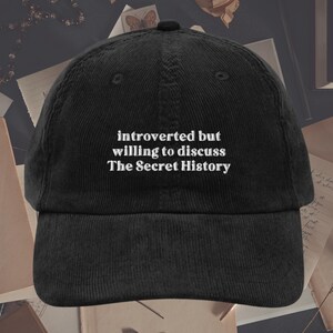 May include: Black corduroy baseball cap with white embroidered text that reads "introverted but willing to discuss The Secret History".