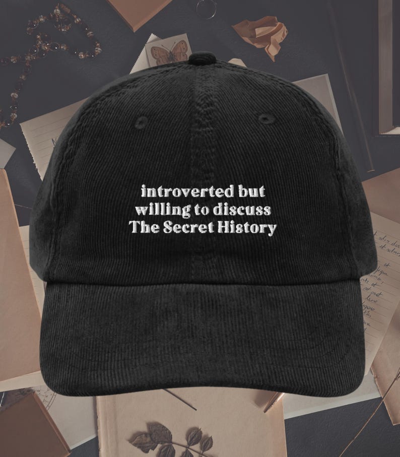 May include: Black corduroy baseball cap with white embroidered text that reads "introverted but willing to discuss The Secret History".