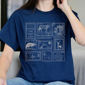 May include: A navy blue t-shirt with a white graphic print of stamps featuring illustrations of a wolf, a castle, a rat, a tree, a deer, a bone, and a dog. The stamps have text that reads "Until the very end", "The Marauders established 1971", "I solemnly swear...", "Rule breakers mischief makers".
