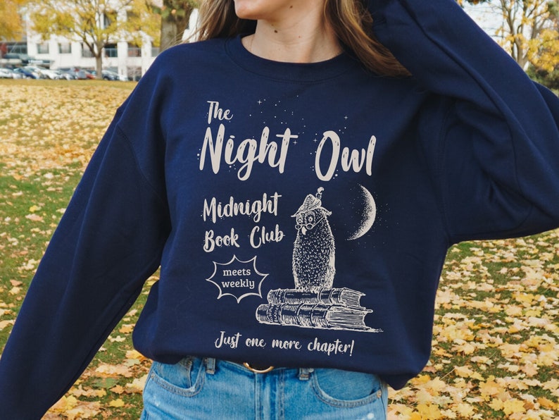 May include: A navy blue sweatshirt with a white graphic design featuring an owl wearing a hat and sitting on a stack of books. The text on the sweatshirt reads "The Night Owl Midnight Book Club meets weekly Just one more chapter!"