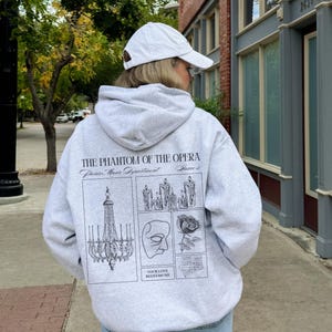 May include: A light gray hoodie with a white baseball cap. The back of the hoodie features a graphic design with the text "THE PHANTOM OF THE OPERA" and illustrations of a chandelier, candles, a mask, and a rose.