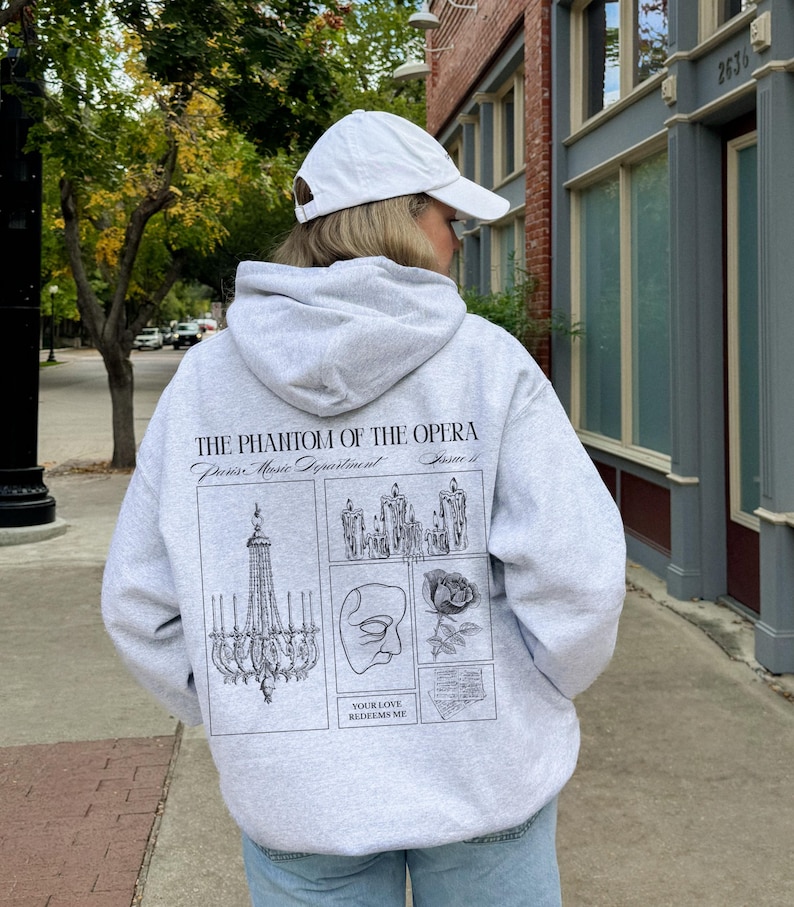 May include: A light gray hoodie with a white baseball cap. The back of the hoodie features a graphic design with the text "THE PHANTOM OF THE OPERA" and illustrations of a chandelier, candles, a mask, and a rose.