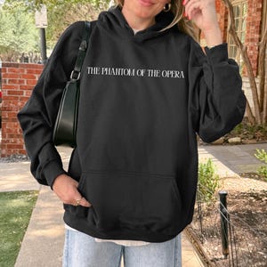 May include: Black hooded sweatshirt with the text "THE PHANTOM OF THE OPERA" in white. The sweatshirt has a front pocket and a drawstring hood. The person is wearing sunglasses and light blue jeans.