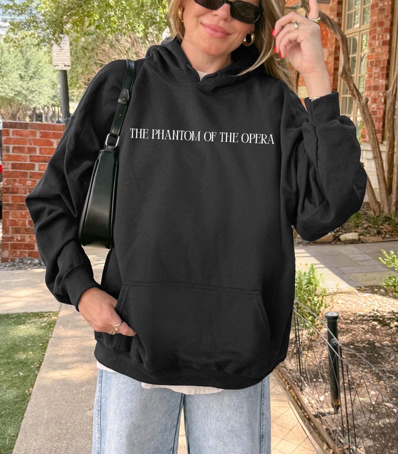 May include: Black hooded sweatshirt with the text "THE PHANTOM OF THE OPERA" in white. The sweatshirt has a front pocket and a drawstring hood. The person is wearing sunglasses and light blue jeans.