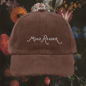 Op de afbeelding: Bruine corduroy baseballcap met de geborduurde tekst "Mood Reader" in witte letters. De cap heeft een gebogen klep en een ronde kroon. De achtergrond toont een bloemstilleven schilderij.