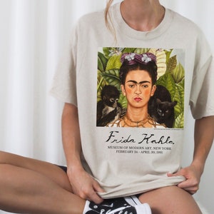 Könnte beinhalten: Ein beiges T-Shirt mit einem Druck von Frida Kahlos Selbstporträt "Die Zwei Fridas." Der Druck enthält den Text "Frida Kahlo. MUSEUM OF MODERN ART, NEW YORK FEBRUARY 24-APRIL 30, 1991."