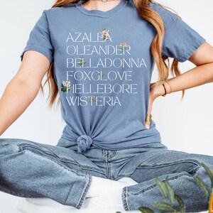 May include: A woman wearing a blue t-shirt with the words "AZALEA, OLEANDER, BELLADONNA, FOXGLOVE, HELLEBORE, WISTERIA" printed on it. She is wearing blue jeans and sitting on a white stool.