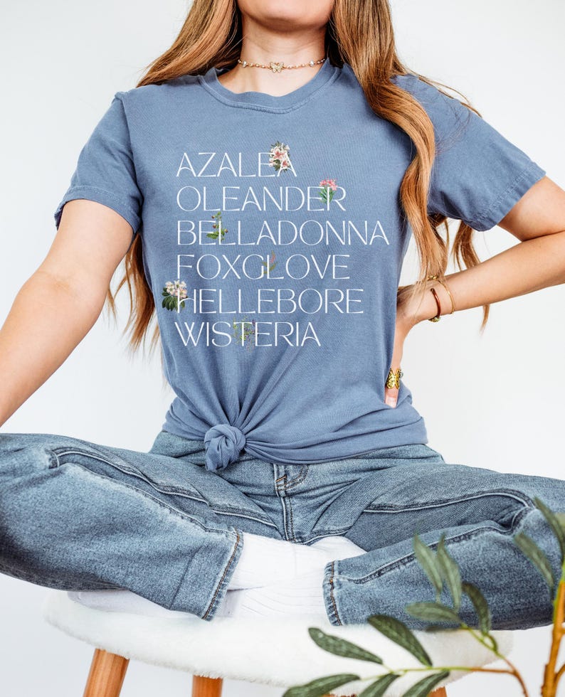 May include: A woman wearing a blue t-shirt with the words "AZALEA, OLEANDER, BELLADONNA, FOXGLOVE, HELLEBORE, WISTERIA" printed on it. She is wearing blue jeans and sitting on a white stool.