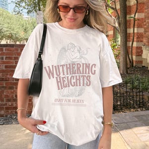 May include: White t-shirt featuring the text "Wuthering Heights" in a pinkish-purple font, along with an illustration of two figures embracing. The phrase "Unfit for Heaven" is printed below the title. The shirt has short sleeves.