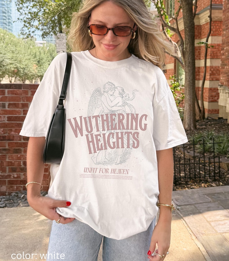 May include: White t-shirt featuring the text "Wuthering Heights" in a pinkish-purple font, along with an illustration of two figures embracing. The phrase "Unfit for Heaven" is printed below the title. The shirt has short sleeves.