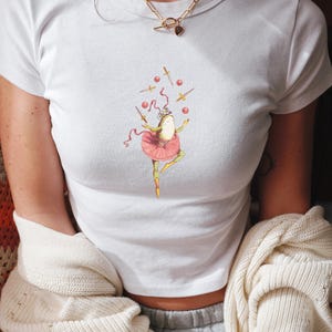 May include: White crop top featuring a whimsical illustration of a frog ballerina juggling swords and red balls. The frog wears a pink tutu and a crown. The image is centered on the shirt.