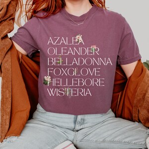 May include: A woman wearing a purple t-shirt with the words "Azalea, Oleander, Belladonna, Foxglove, Hellebore, Wisteria" printed on it. Each word is accompanied by a small floral illustration.