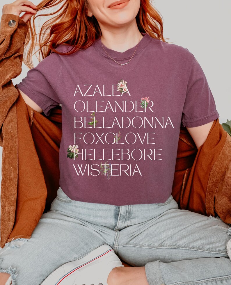 May include: A woman wearing a purple t-shirt with the words "Azalea, Oleander, Belladonna, Foxglove, Hellebore, Wisteria" printed on it. Each word is accompanied by a small floral illustration.