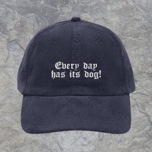 May include: A dark navy blue corduroy baseball cap featuring white embroidered text on the front panel that reads, Every day has its dog! The cap has a structured crown, a curved brim, and a button at the top center of the fabric dome.
