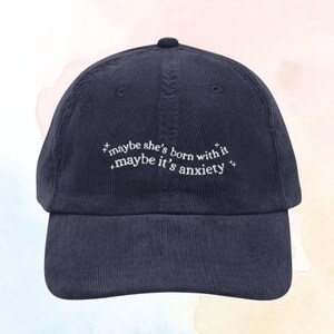 May include: A navy blue corduroy baseball cap with white embroidered text that reads "maybe she's born with it maybe it's anxiety".