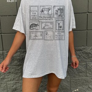 May include: A gray oversized t-shirt with a graphic print of black and white stamps. The stamps feature images of a wolf, a castle, a deer, a rat, a dog, and a log. The text on the stamps reads "Until the very end", "The Marauders established 1971", "Rule Breakers Mischief Makers".