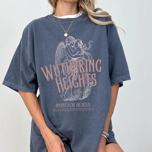 May include: Denim blue t-shirt featuring the text "Wuthering Heights" in a pink hue, along with an illustration of two figures embracing. The phrase "Unfit for Heaven" is printed below the illustration. The shirt has short sleeves.