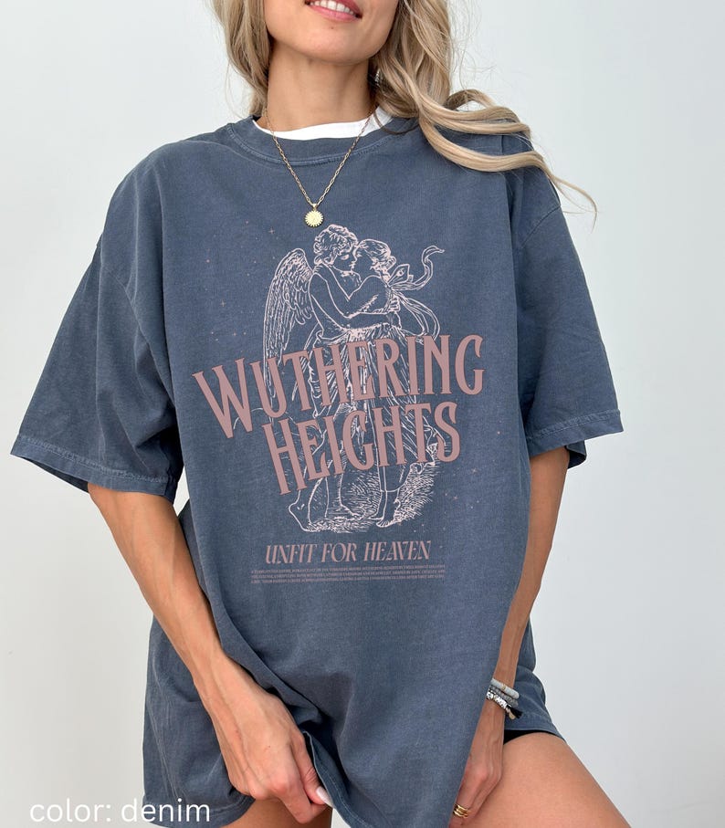 May include: Denim blue t-shirt featuring the text "Wuthering Heights" in a pink hue, along with an illustration of two figures embracing. The phrase "Unfit for Heaven" is printed below the illustration. The shirt has short sleeves.