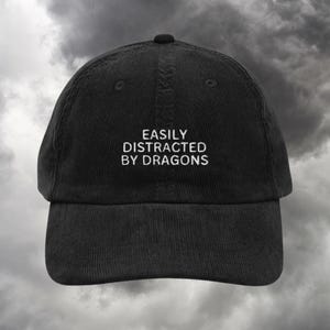 以下が含まれることがあります： 「Easily Distracted by Dragons」という白い刺繍が施された黒いコーデュロイ製のベースボールキャップです。