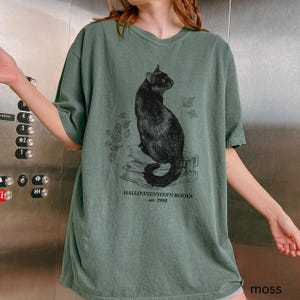 Op de afbeelding: Groen t-shirt met een zwarte kat illustratie op boeken en de tekst "Halloweentown Books est. 1998". Het shirt heeft korte mouwen en een losse pasvorm.