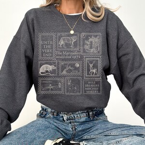 K&ouml;nnte beinhalten: Ein dunkelgraues Sweatshirt mit einem grafischen Druck von Briefmarken mit verschiedenen Bildern, darunter ein Wolf, eine Ratte, ein Reh und ein Schloss. Der Text auf den Briefmarken lautet "Until the very end", "The Marauders established 1971" und "Rule Breakers Mischief Makers".